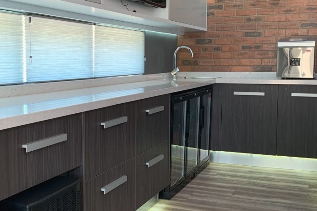 Modern kitchen area featuring sleek dark cabinetry, stone countertops, an exposed brick feature wall, and integrated under-counter beverage refrigeration with ambient LED lighting.