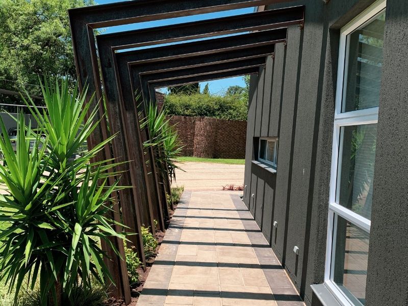 A modern exterior walkway featuring a striking steel pergola structure, paved path, and well-maintained textured wall finish along the side of the house.