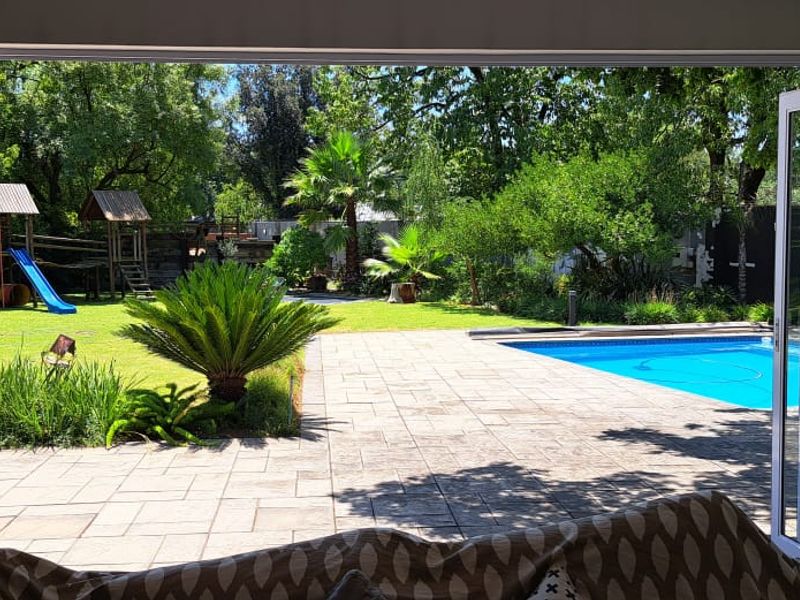 View from an indoor living space through large folding glass doors onto a paved patio, swimming pool, and landscaped garden area.