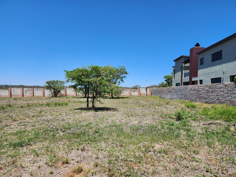 A view of the vacant residential land featuring a stone-clad boundary wall and established trees, adjacent to an existing multi-story residential structure.