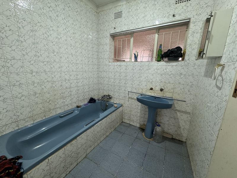 A dated bathroom featuring a blue bathtub and matching pedestal sink, fully tiled walls, and a window with security bars.