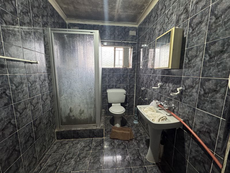 A dated bathroom featuring dark marble-effect wall and floor tiles, a shower enclosure, pedestal basin, and toilet.