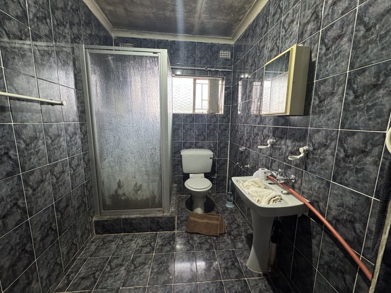 A dated bathroom featuring dark tiled walls and floors, a shower enclosure, pedestal basin, and toilet, requiring modernization and cosmetic updates.