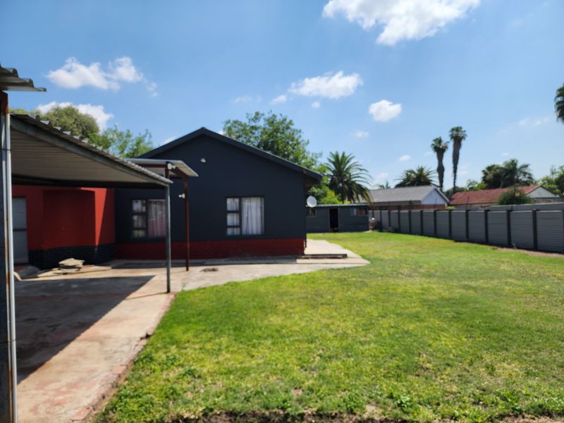 Rear view of the property featuring a covered patio area, concrete paving, and a spacious garden enclosed by metal perimeter fencing.
