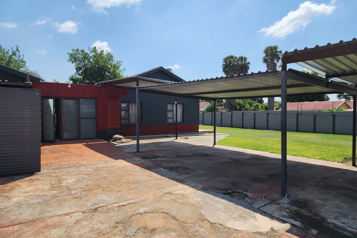 Rear exterior view featuring a large concrete driveway, a metal carport structure, and a spacious lawn enclosed by metal fencing.