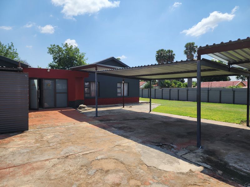 Rear exterior view featuring a large concrete driveway, a metal carport structure, and a spacious lawn enclosed by metal fencing.