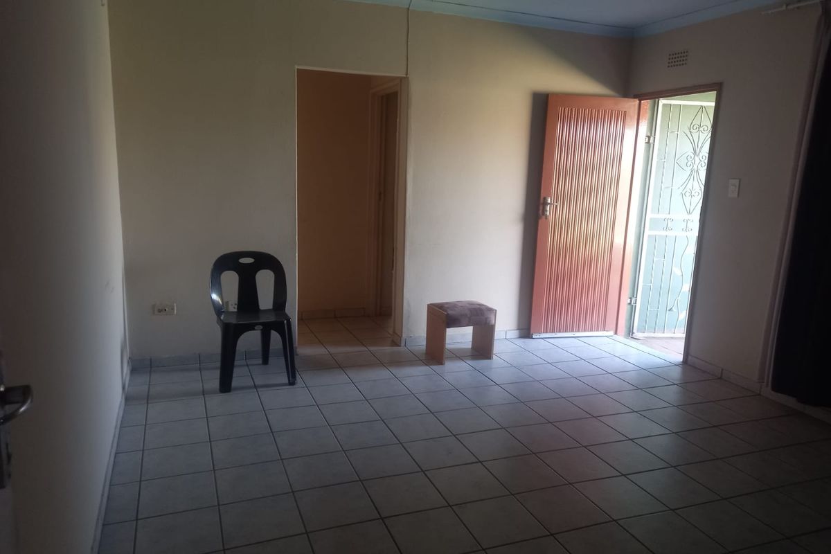 A sparsely furnished living room featuring tiled flooring, a single entrance door with security gate, and access to an adjacent hallway.