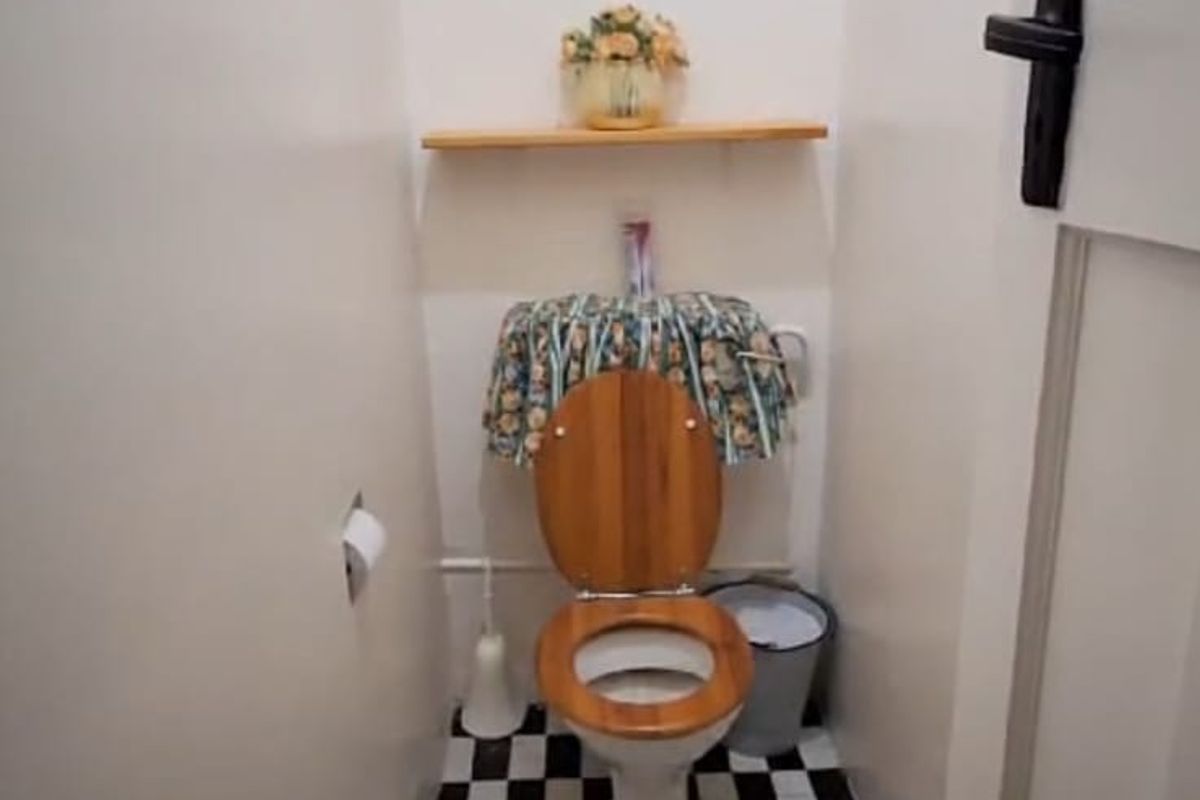 A compact guest toilet featuring a wooden seat, checkered floor tiling, and a small wooden-framed window.