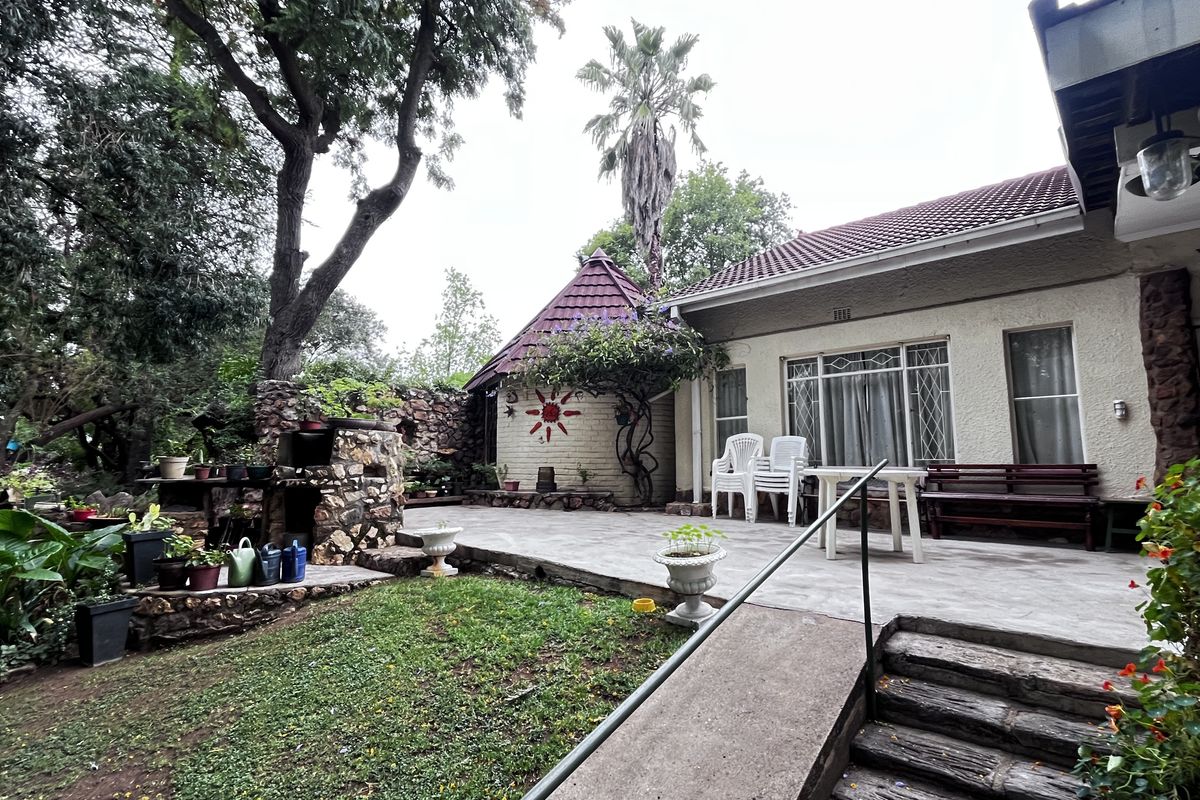 A spacious rear patio area featuring a stone-built outdoor braai, established garden surroundings, and direct access to the main dwelling.