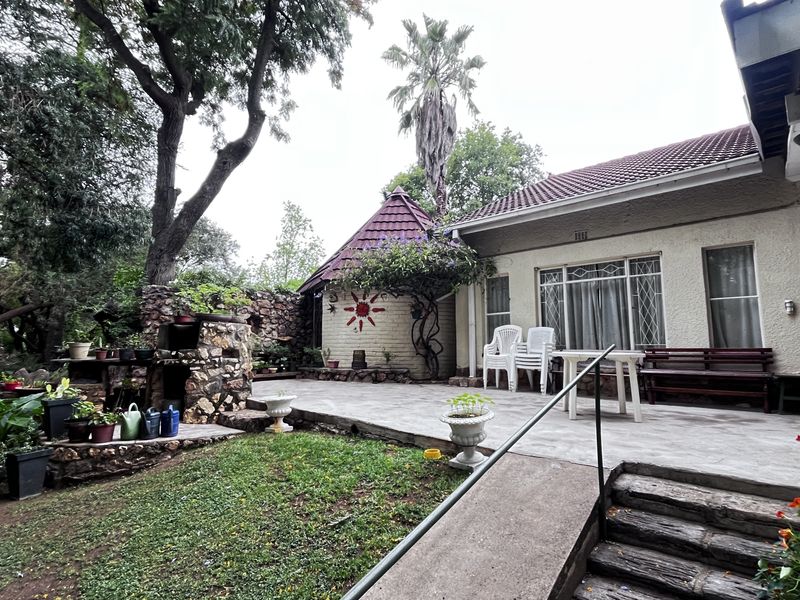 A spacious rear patio area featuring a stone-built outdoor braai, established garden surroundings, and direct access to the main dwelling.