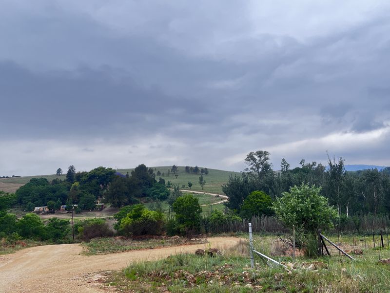 A scenic view of the rural landscape and surrounding small holding terrain, featuring a gravel access road and natural vegetation under a cloudy sky.