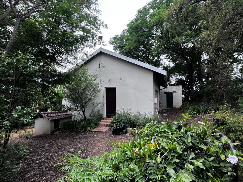 Rear view of a small holding dwelling featuring a simple gable roof design, set within a mature, wooded garden environment.