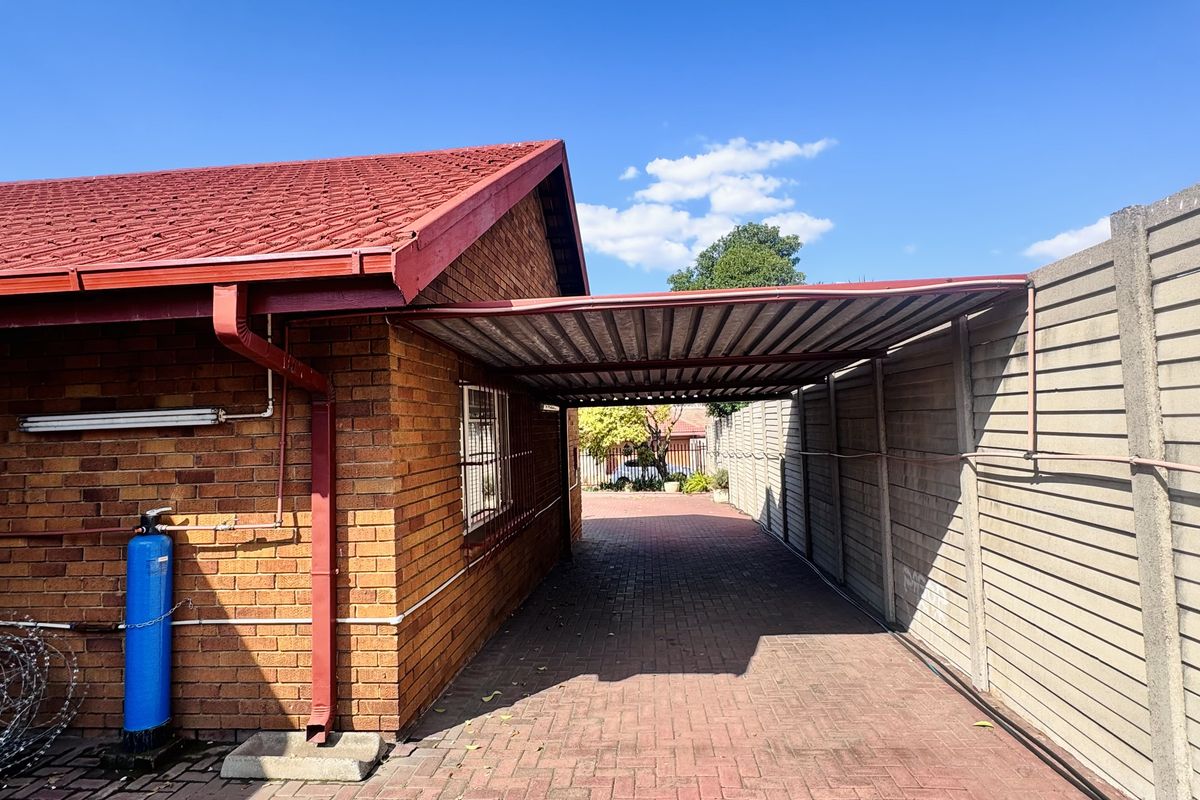 A paved driveway area featuring a covered carport structure adjacent to the brick house exterior and perimeter boundary wall.