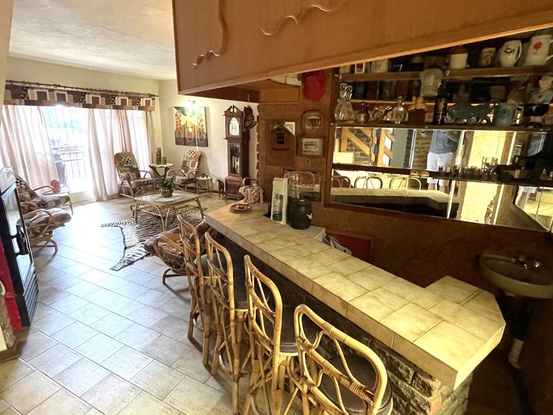 A dated home bar area featuring a tiled counter, built-in sink, and mirrored wall, adjacent to an open-plan living space with tiled flooring.