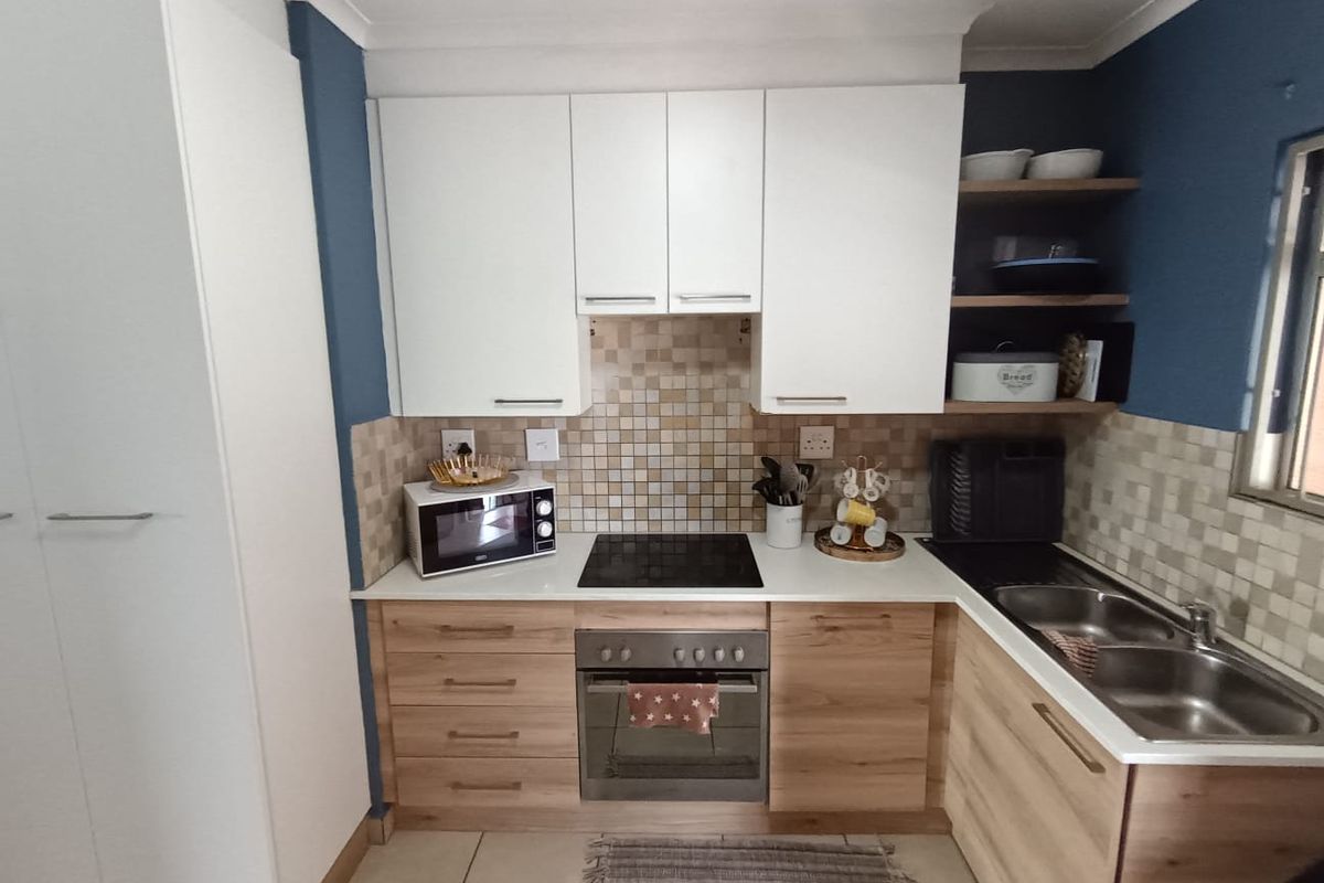 A compact, well-maintained kitchen featuring modern white and wood-grain cabinetry, a mosaic tile backsplash, integrated oven and hob, and a double stainless steel sink.