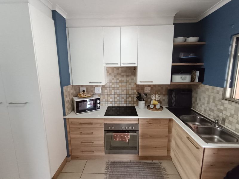 A compact, well-maintained kitchen featuring modern white and wood-grain cabinetry, a mosaic tile backsplash, integrated oven and hob, and a double stainless steel sink.