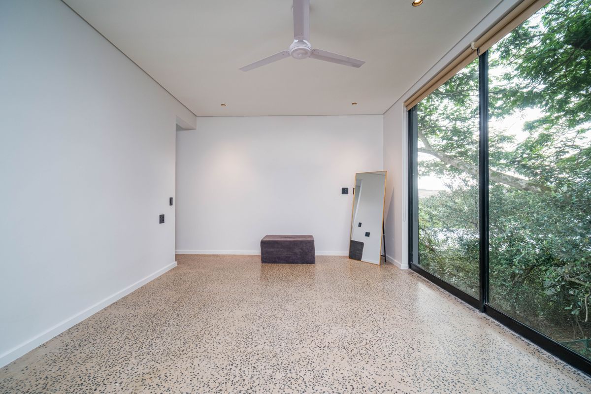 A bright, modern bedroom featuring polished concrete flooring, a ceiling fan, and large floor-to-ceiling sliding glass doors overlooking lush greenery.