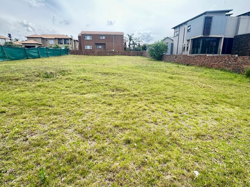 A vacant residential plot featuring a grassed surface, enclosed by brick boundary walls and green mesh fencing in a developed neighborhood.