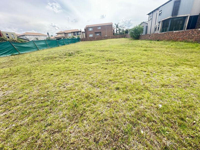 A vacant residential plot featuring a grassed surface, enclosed by a brick boundary wall and temporary green shade cloth fencing.