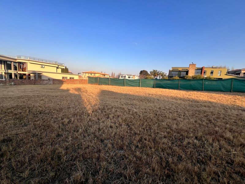 A vacant residential plot featuring dry grass and perimeter fencing, situated in a developed neighborhood with surrounding modern homes.