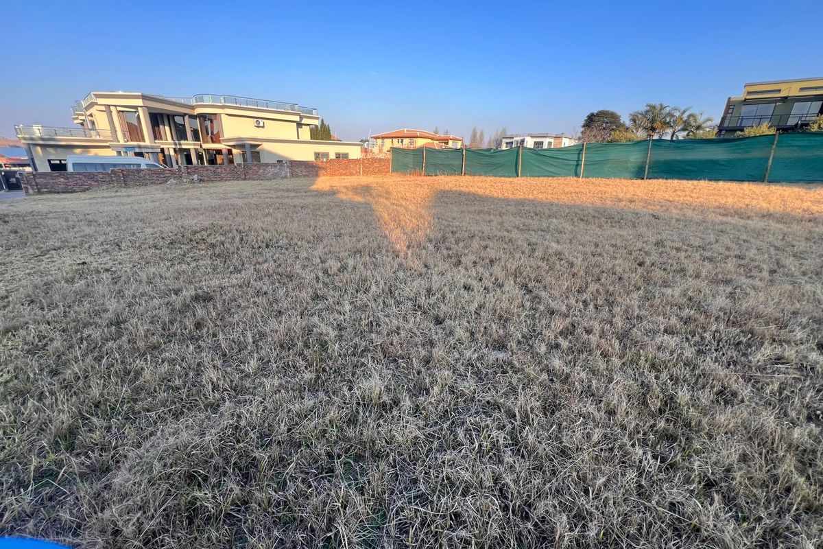 A vacant residential plot featuring dry grass, enclosed by a boundary wall and privacy screening in a developed neighborhood.