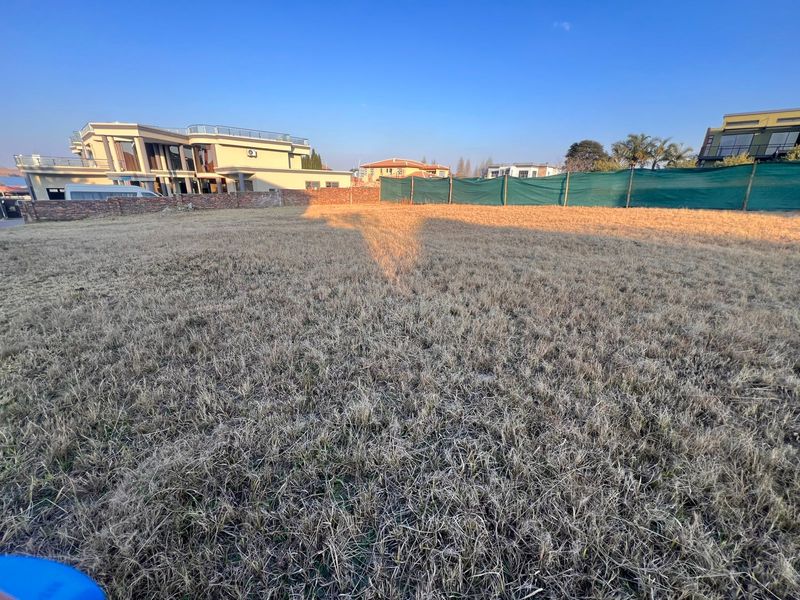 A vacant residential plot featuring dry grass, enclosed by a boundary wall and privacy screening in a developed neighborhood.