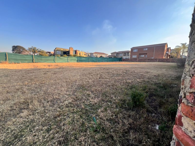 A vacant residential plot featuring dry grass and perimeter fencing with green shade cloth screening, situated in a developed neighborhood.