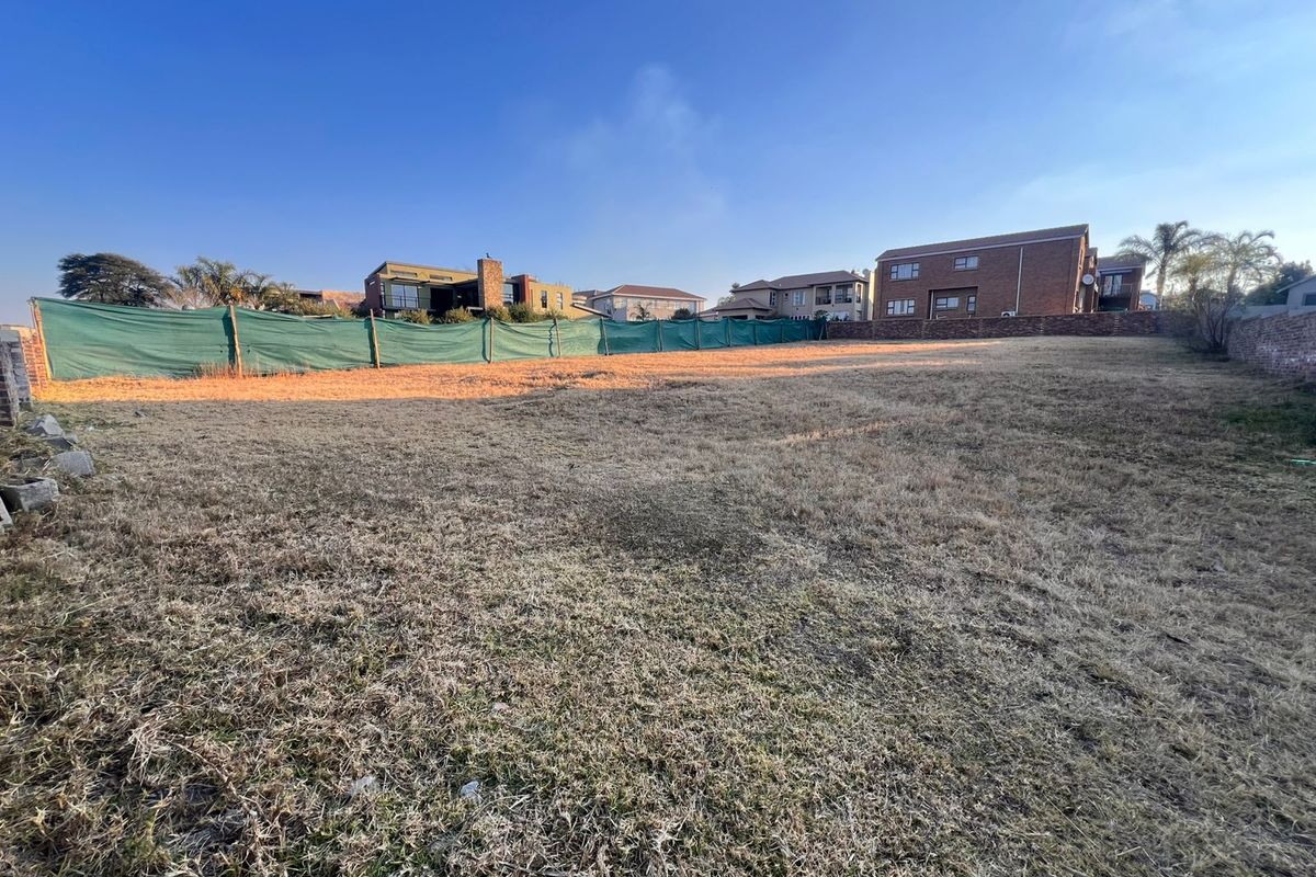 A vacant residential plot featuring dry grass, enclosed by a brick boundary wall and green shade cloth fencing in a developed neighborhood.