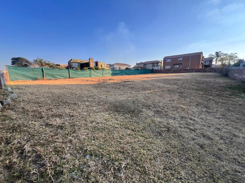 A vacant residential plot featuring dry grass, enclosed by a brick boundary wall and green shade cloth fencing in a developed neighborhood.