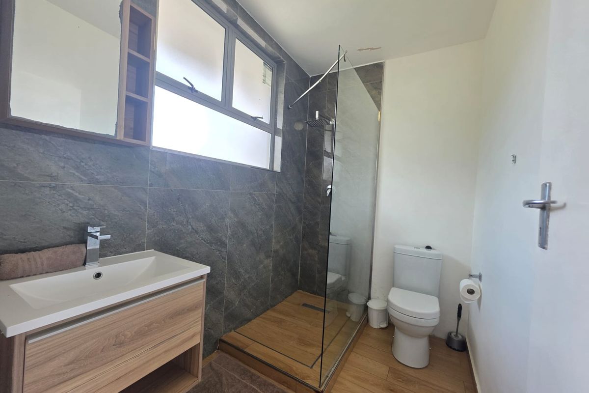 Modern bathroom featuring a glass-enclosed shower, wood-look floor tiles, dark stone-effect wall tiles, and a contemporary vanity unit with a basin.