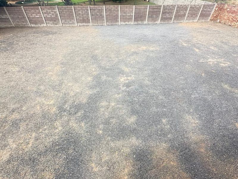 A spacious, level vacant residential plot featuring a gravel surface and enclosed by perimeter precast concrete walling and mature trees.
