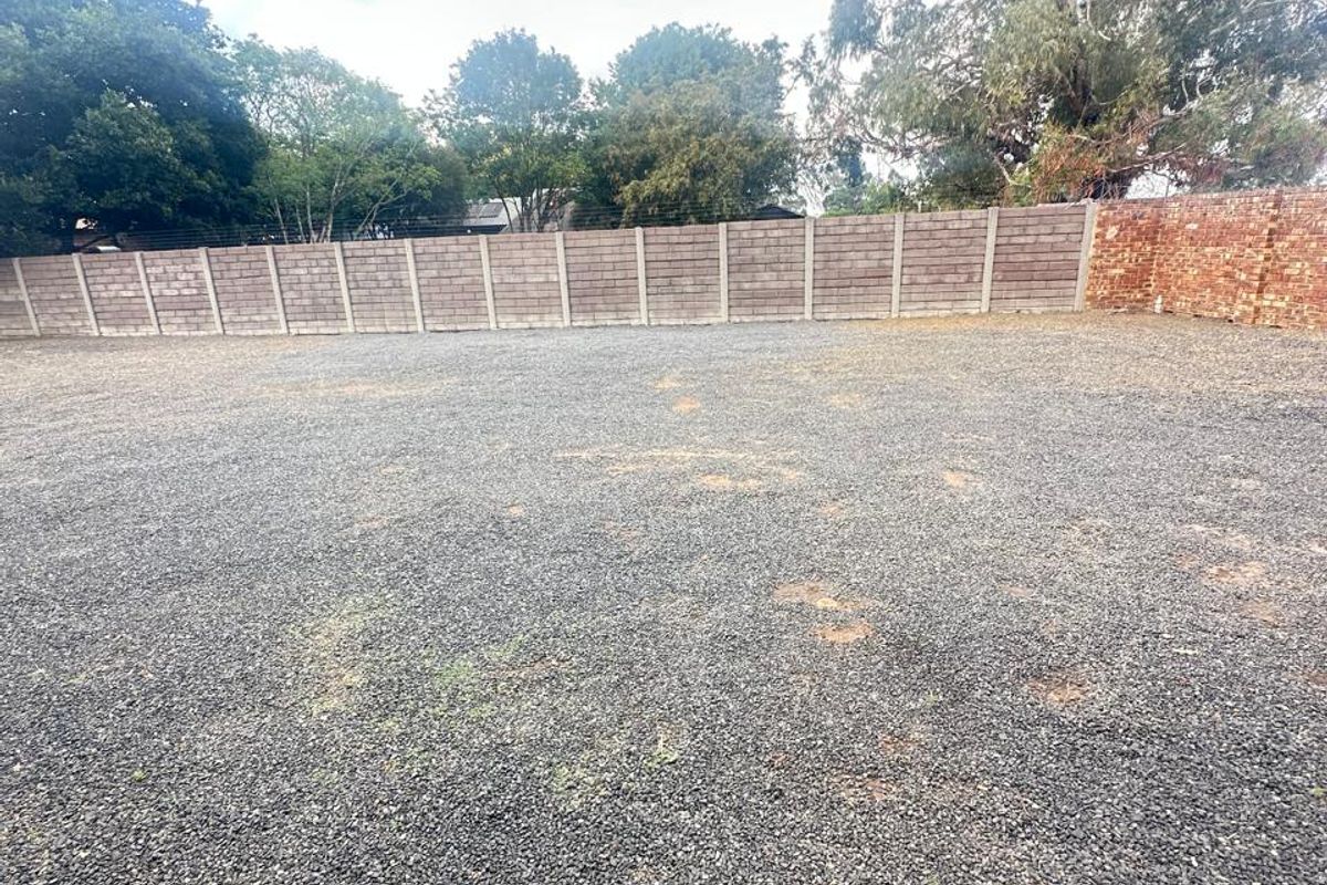 A vacant residential plot featuring a gravel surface, enclosed by perimeter concrete block and brick boundary walls with mature trees in the background.
