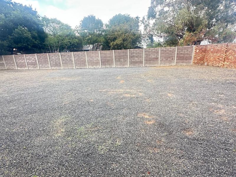 A vacant residential plot featuring a gravel surface, enclosed by perimeter concrete block and brick boundary walls with mature trees in the background.