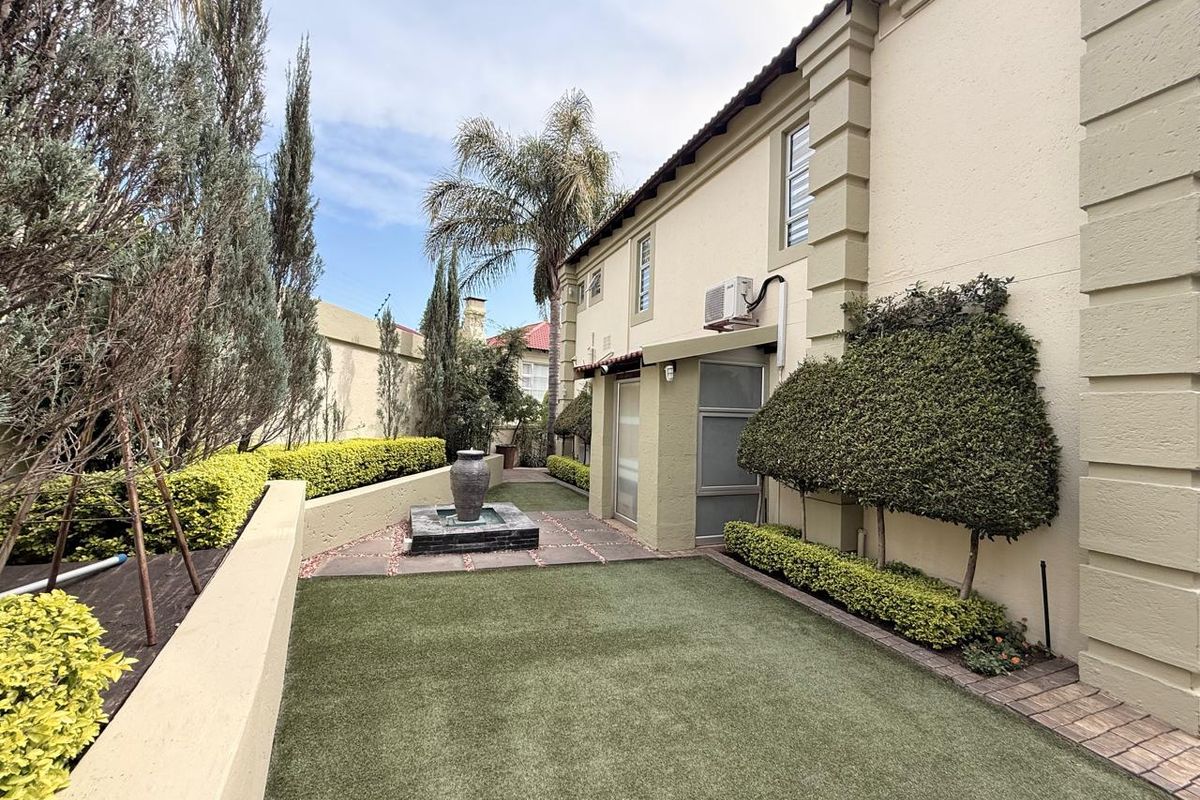 A well-maintained rear garden featuring artificial turf, a decorative water feature, manicured hedges, and a paved walkway alongside the townhouse exterior.