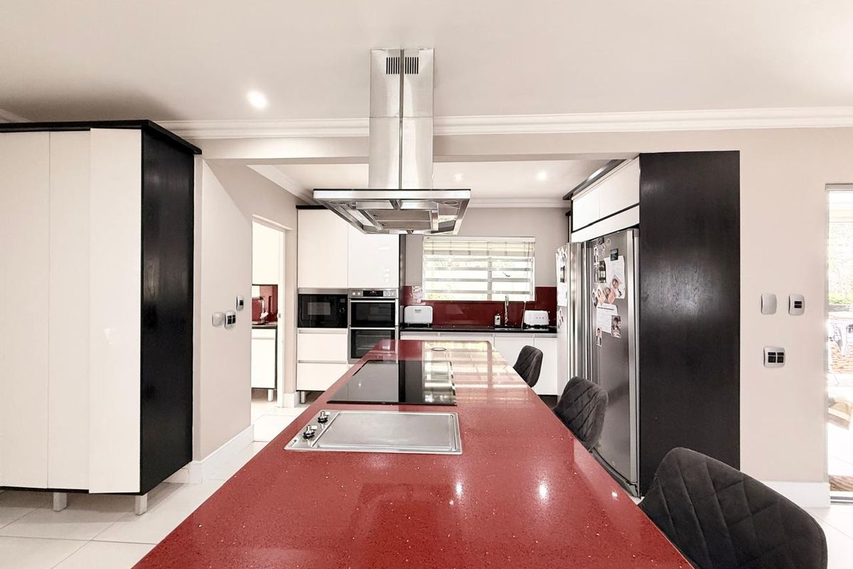 Modern kitchen featuring a large red quartz island with integrated hob, stainless steel extractor fan, built-in ovens, and sleek high-contrast cabinetry.