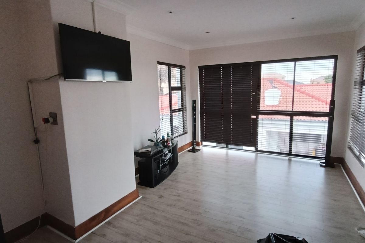 A bright living room featuring light-toned laminate flooring, large windows with fitted blinds, and recessed ceiling lighting.