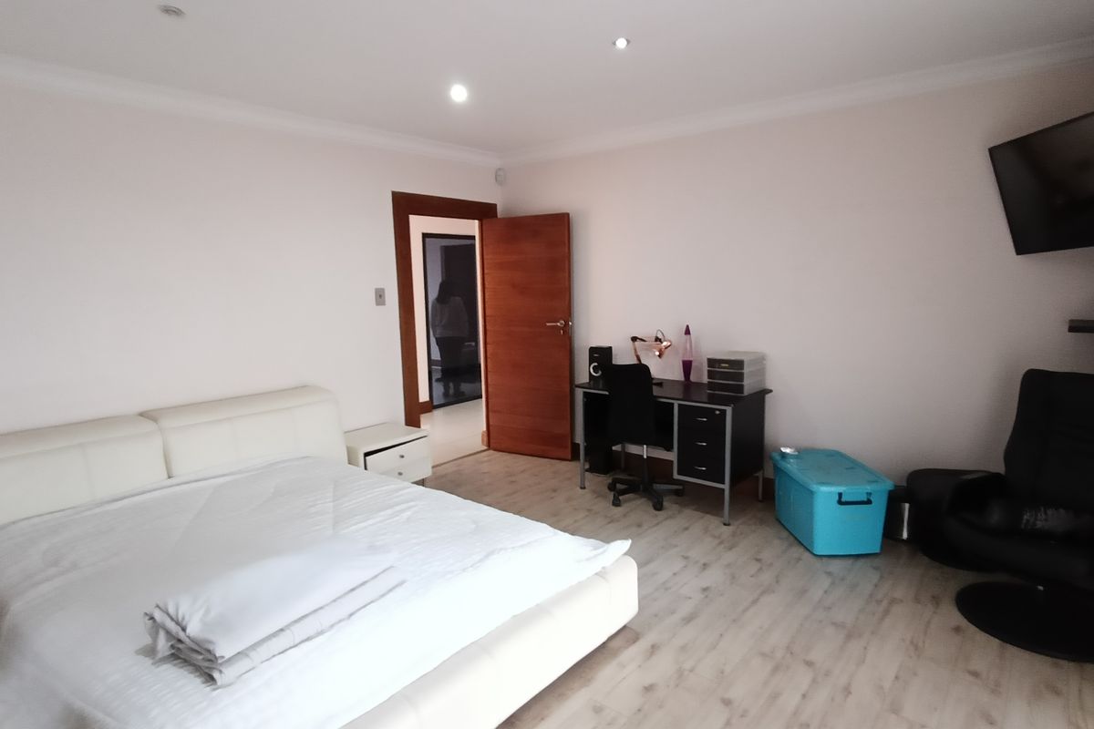 A spacious bedroom featuring light-toned laminate flooring, recessed ceiling lighting, and a solid timber door leading to an adjacent hallway.