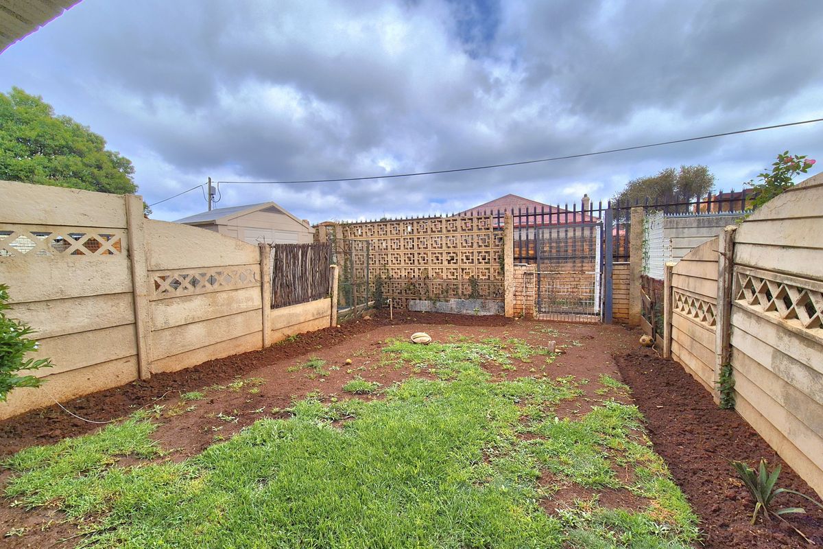 A rear garden area featuring precast concrete boundary walls, a metal security gate, and an open lawn space with exposed soil patches.