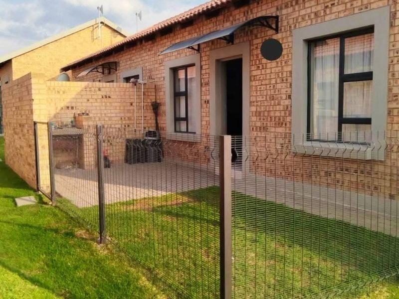 Rear exterior view of a townhouse featuring a brick facade, paved patio area with a built-in braai, and enclosed garden space with wire fencing.