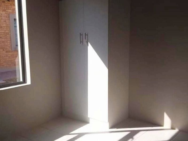 A simple, empty bedroom featuring tiled flooring and a built-in wardrobe, receiving natural light through a side window.