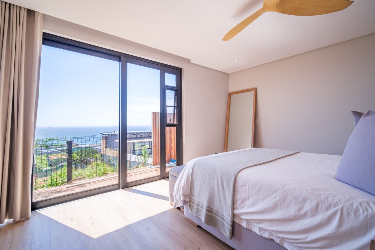 A bright, modern bedroom featuring light wood flooring, a ceiling fan, and large sliding glass doors that open to a scenic coastal view.