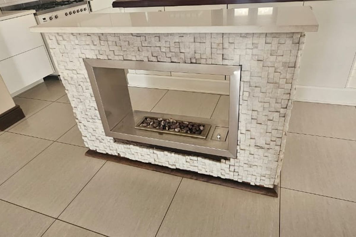 A modern kitchen area featuring a unique central island with a built-in fireplace, stone mosaic detailing, and neutral ceramic floor tiles.