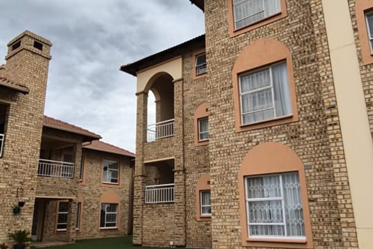 Exterior view of a multi-story brick apartment complex featuring arched balconies, tiled roofing, and a paved driveway area.