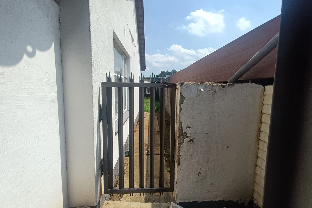A side exterior passage featuring a metal security gate, concrete pathway, and boundary wall with visible signs of weathering and paint deterioration.