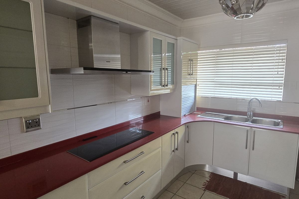 A functional kitchen featuring white cabinetry, a bold red countertop, integrated hob, stainless steel extractor fan, and a double sink with tiled finishes.