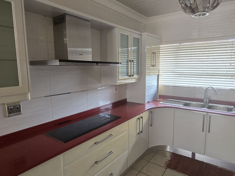 A functional kitchen featuring white cabinetry, a bold red countertop, integrated hob, stainless steel extractor fan, and a double sink with tiled finishes.