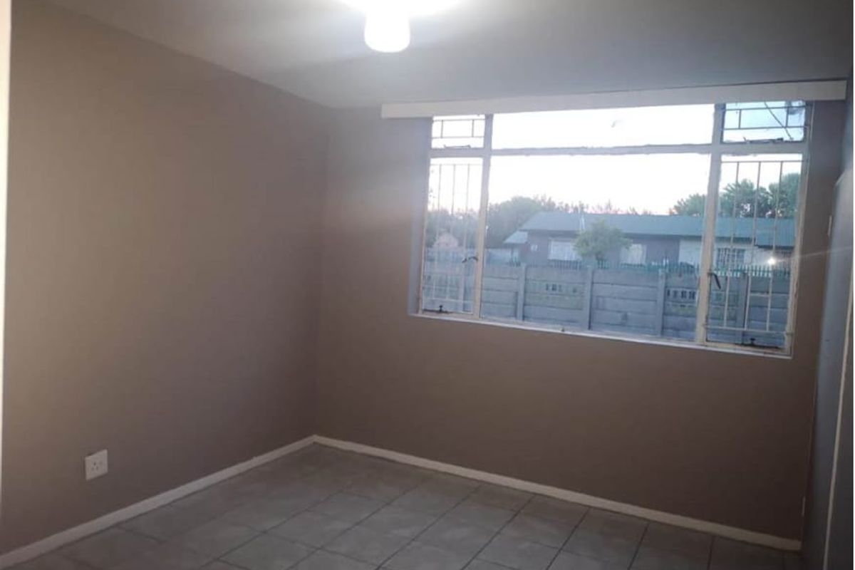 A simple, empty bedroom featuring tiled flooring, neutral walls, and a large window with security bars.