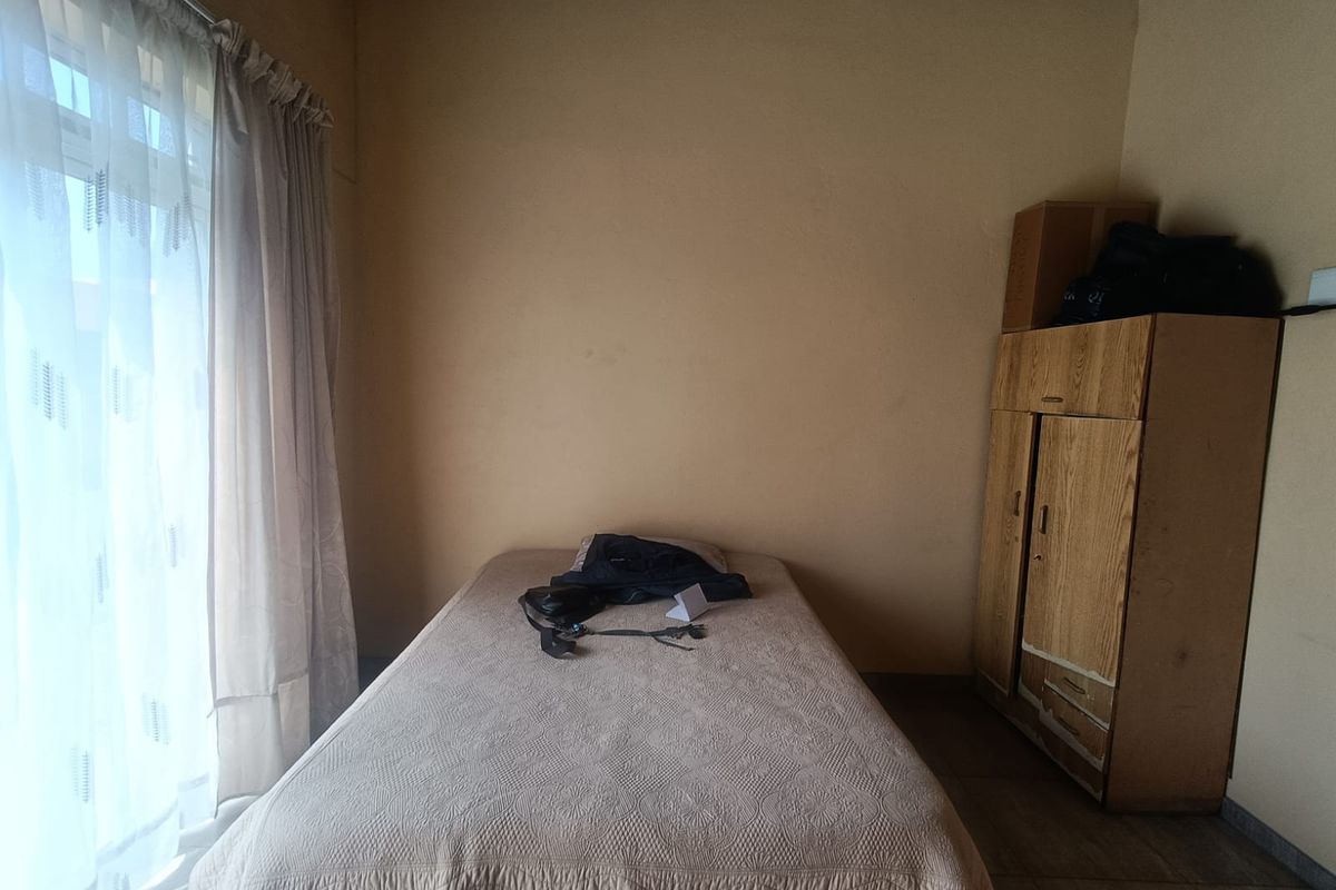 A bedroom featuring tiled flooring and a large window with curtains, showing signs of wear and an unfinished ceiling.