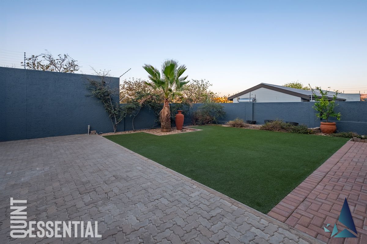 A well-maintained rear garden featuring a paved driveway area, artificial lawn, and high perimeter boundary walls with electric fencing.