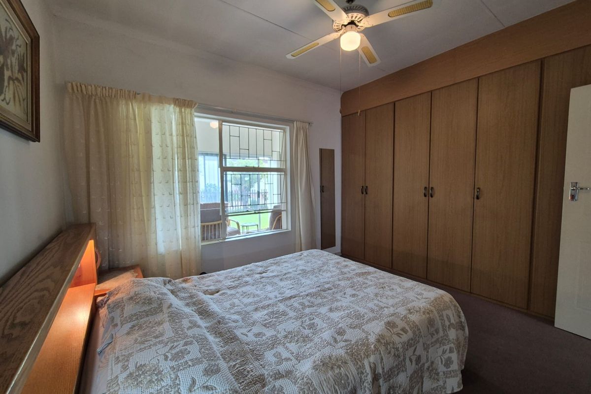 A bedroom featuring built-in wooden cupboards, a ceiling fan, and a window with security bars overlooking the garden.